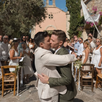 Gay couple kissing at a destination wedding in Greece, both in wool suits—one olive green, one beige—on a hot sunny day.
