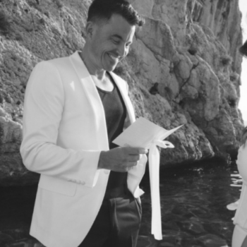 Groom in a black wool suit trousers and ivory tuxedo jacket, looking down at vows during a seaside wedding in Italy.