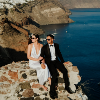 Bride and groom sitting on a rock with Santorini cliffs and the sea in the background, groom in a black wool dinner suit for a Greek islands elopement.
