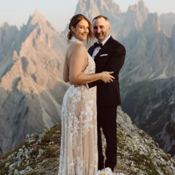 Bride and groom standing atop a mountain in the Italian Dolomites, groom in a black wool dinner suit, dramatic alpine view behind them.